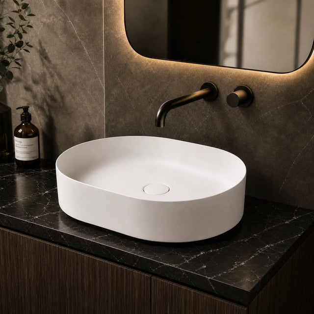 White oval sink on a dark marble countertop with a modern mirror and decorative bottles.